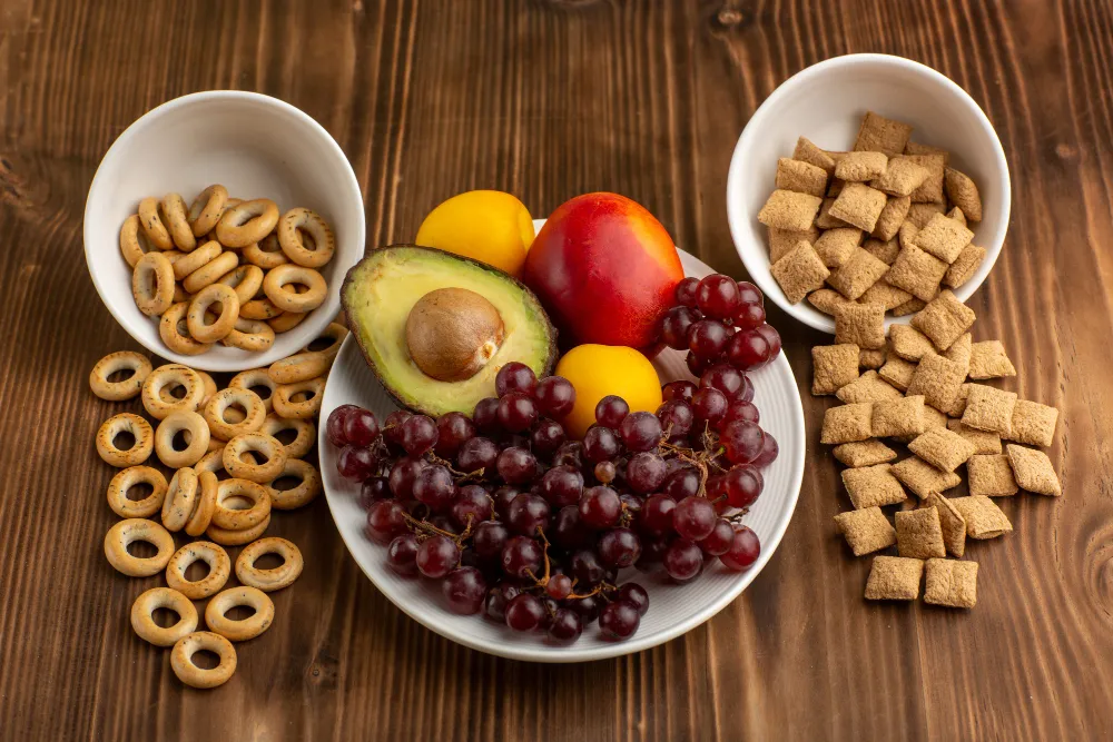 Low-calorie snack spread with red grapes, avocado, fresh stone fruit, crunchy cereal squares, and baked mini rings